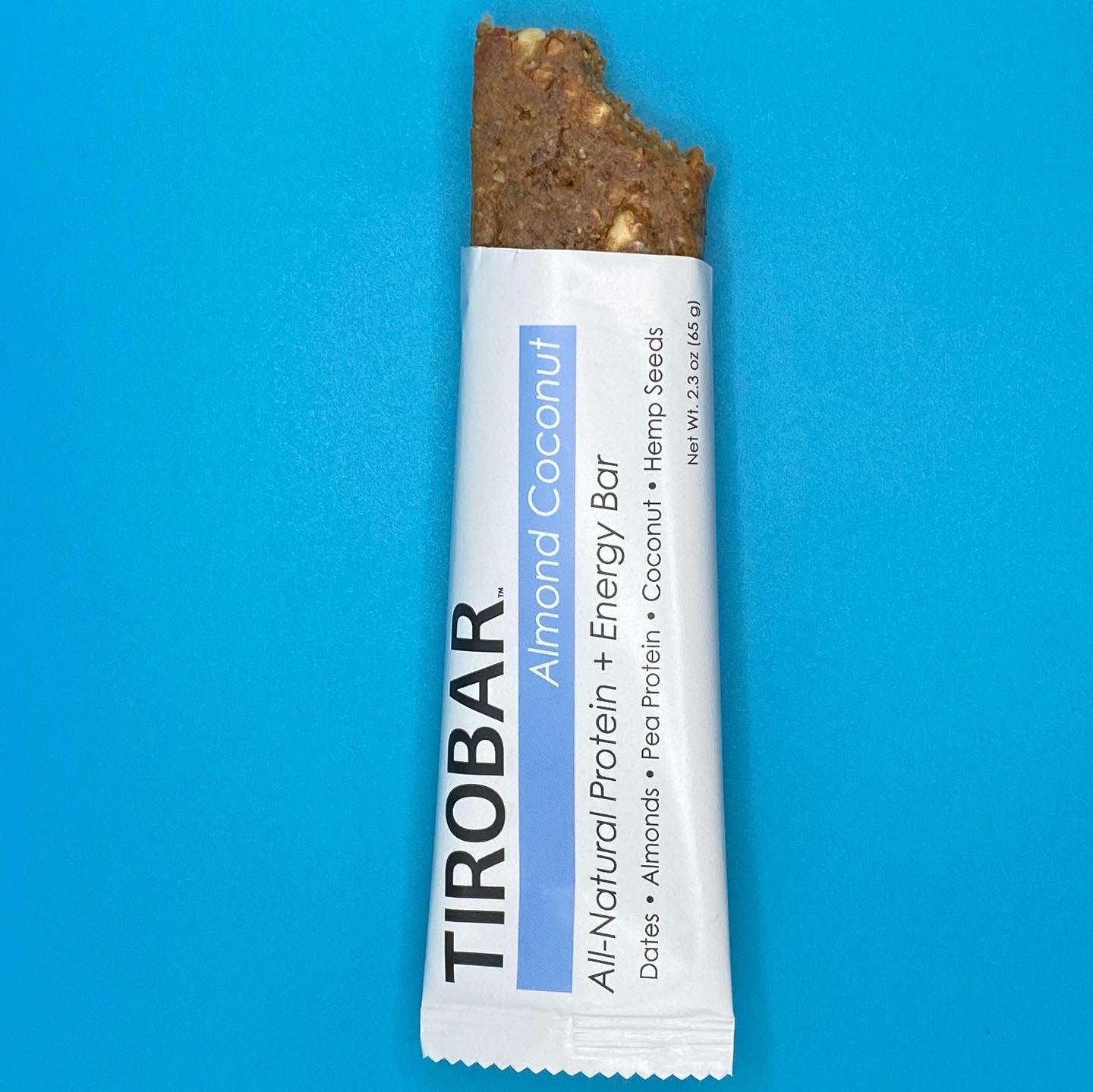An almond coconut TIROBAR, a vegan and all natural protein bar, sits in a white wrapper against a blue background. The bar is visible from the top, showing the almonds, coconuts, and consistency of the protein bar. 