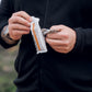 A man in a black sweatshirt opens an all natural, vegan salted peanut TIROBAR protein bar. The wrapper of the bar is visible and the man's hand is holding the bar. The background is blurred.
