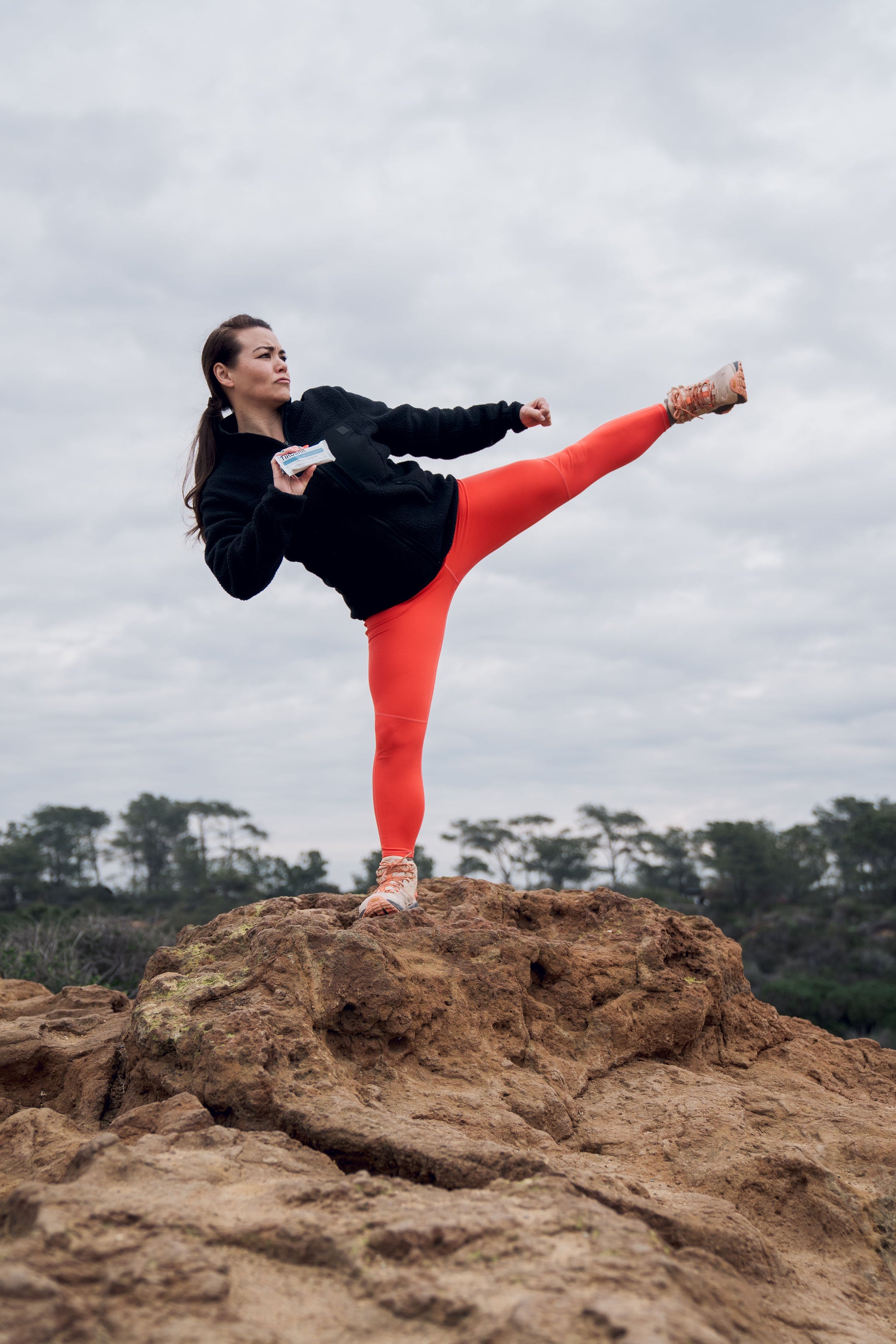A woman performs a kick while working out on top of a mountain, holding an almond coconut TIROBAR, an all natural and vegan protein bar. The woman wears workout clothes and stands on a rocky surface overlooking a scenic view.