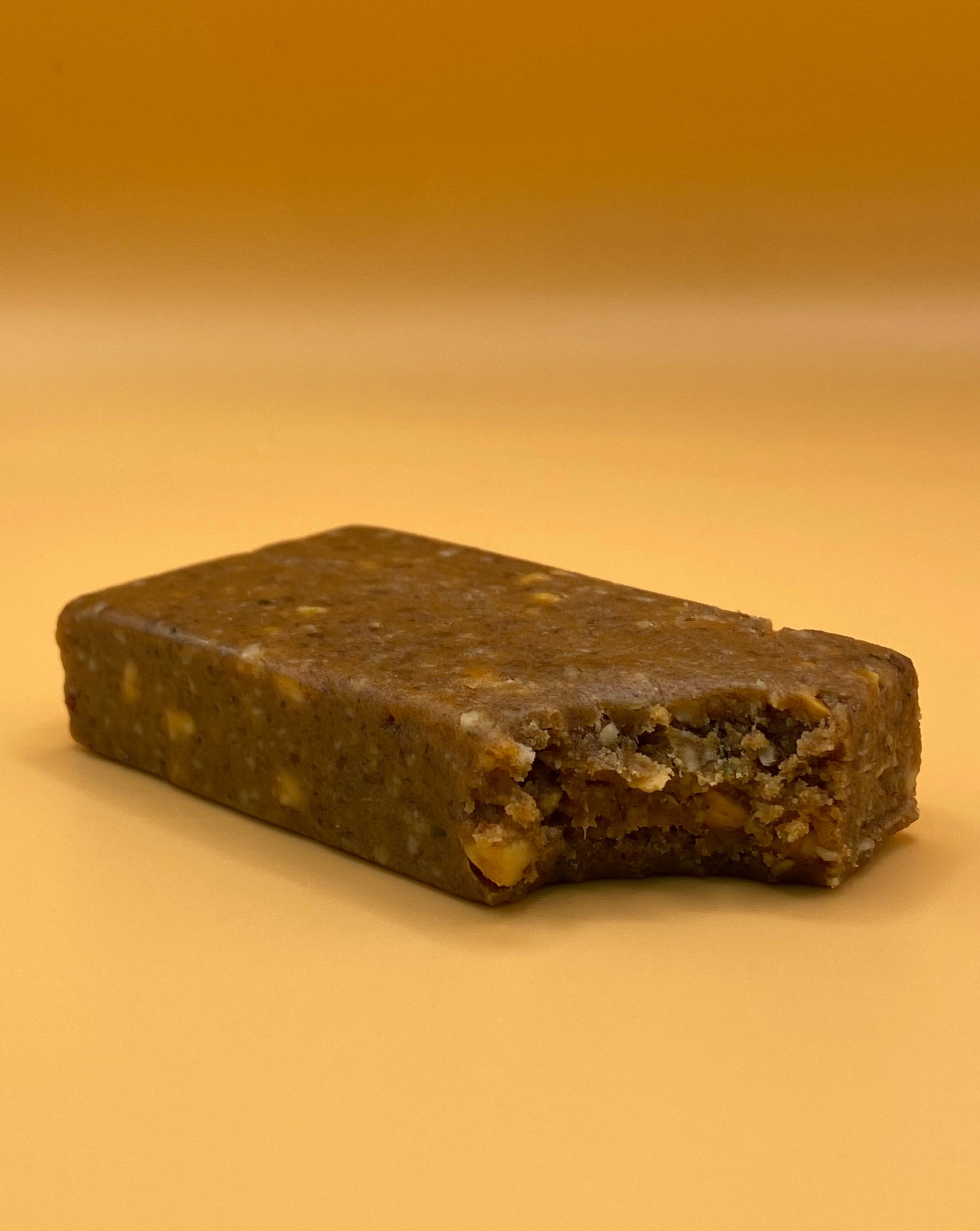 A salted peanut TIROBAR, a vegan and all natural protein bar, is shown bitten with peanuts visible in the filling. The bar is placed against an orange background.