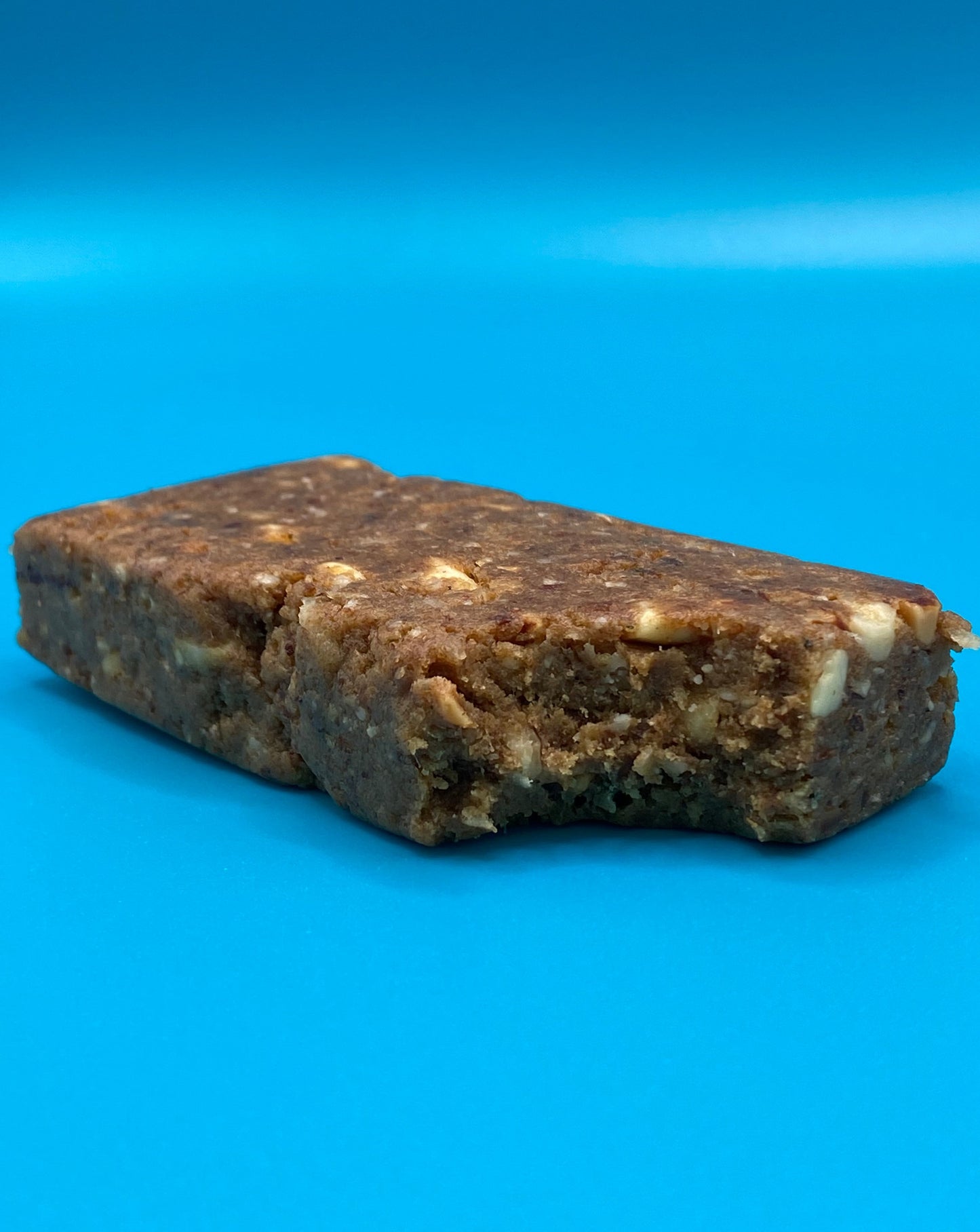 An almond coconut TIROBAR, a vegan and all natural protein bar, is shown bitten with coconut flakes and almonds visible in the filling. The bar is placed against a blue background.