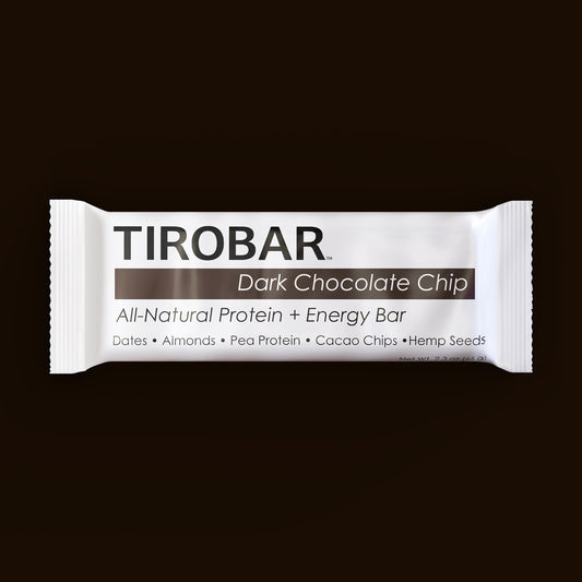 A dark chocolate chip TIROBAR, a vegan and all natural protein bar, sits in a white wrapper against a brown background. The bar is visible from the top with view of the ingredients.